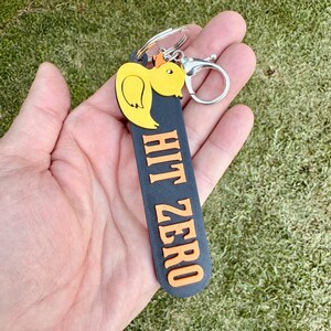 May include: Black and orange keychain with a yellow rubber duck charm. The keychain has the text "HIT ZERO" printed on it.