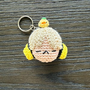 May include: A handmade crochet keychain featuring a cartoon character with closed eyes, light skin, and blonde hair. The keychain has yellow accents and a small yellow duck on top. The keychain is attached to a silver metal ring.