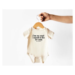 Pode incluir: Um macacão de bebê de cor creme com mangas curtas e um gráfico preto que diz "i'm so cool i used to be fr*zen" com um símbolo de floco de neve e o texto "#irl baby"