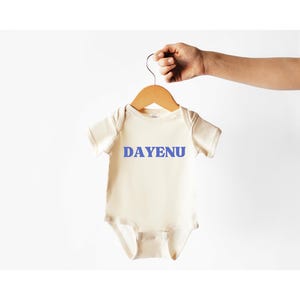 May include: A cream-coloured baby onesie with short sleeves and a snap closure. The onesie has the word "DAYENU" printed in blue on the front.