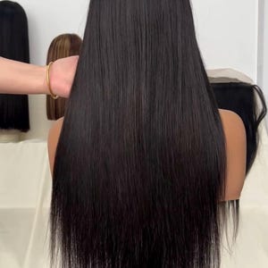 Pode incluir: Uma peruca preta com cabelo comprido e liso é exibida em uma cabeça de manequim. O cabelo é brilhante e cai pelas costas. Outras perucas são visíveis ao fundo. A peruca é provavelmente para modelar e usar.