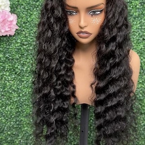 May include: A long, black, curly wig styled on a mannequin head. The wig features a natural-looking hairline and voluminous waves. The mannequin is mounted on a black stand against a green backdrop.