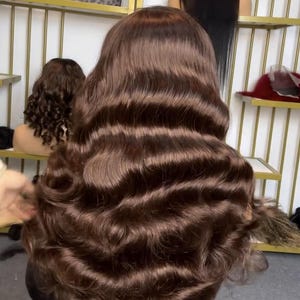 May include: A brown, wavy wig on a mannequin head. The wig has long, flowing hair with a deep brown colour. Other wigs are visible in the background, displayed on shelves. The hair is styled in loose waves.