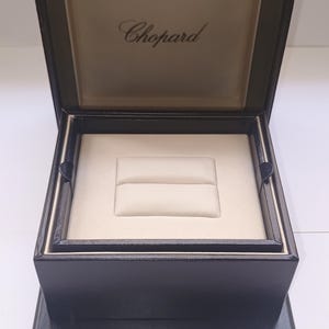 Può includere: Un portagioie nero di forma quadrata con coperchio incernierato aperto che rivela un interno color crema. L'interno del coperchio presenta il testo color oro Chopard. La base contiene un inserto centrale imbottito color crema.