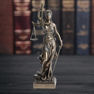 May include: Bronze statue of Lady Justice, blindfolded, holding scales and a sword. The statue is set against a backdrop of antique books, suggesting a theme of law and justice. The figure is detailed and stands on a base.