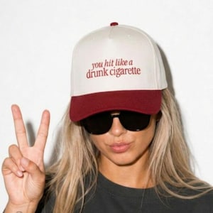May include: A baseball cap with a cream-colored crown and a dark red brim. The front of the cap features the text you hit like a drunk cigarette embroidered in dark red lettering. The cap has a structured front panel and a button on the top.