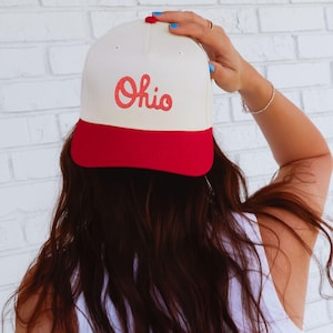 May include: A cream and red baseball cap with the word "Ohio" in red script. The cap has a red peak and a cream crown. The person is wearing a white vest top.