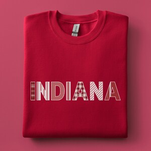 May include: A red sweatshirt featuring the word "INDIANA" in white, patterned lettering. The patterns include stripes, polka dots, and plaid. The sweatshirt is folded on a red surface.