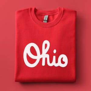 May include: A folded red sweatshirt with the white word "Ohio" in a cursive font. The sweatshirt is made of a soft material and is displayed on a red background. The Gildan tag is visible.
