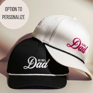 May include: Two baseball caps, one black and one white, are stacked. The black cap has white embroidered text that says "Girl Dad". The white cap has pink embroidered text that says "Girl Dad". Both caps have a white rope detail along the brim.