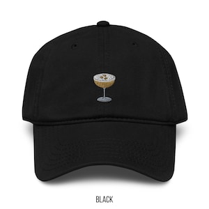 May include: Black baseball cap with an embroidered cocktail glass design. The cocktail glass is gold with a silver stem and contains a brown liquid with white foam and brown specks. The word "BLACK" is printed at the bottom.