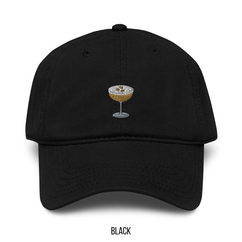 May include: Black baseball cap with an embroidered cocktail glass design. The cocktail glass is gold with a silver stem and contains a brown liquid with white foam and brown specks. The word "BLACK" is printed at the bottom.