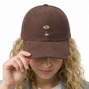 May include: A brown corduroy baseball cap with a small embroidered design of a cocktail glass. The cap has a curved brim and a button on top. The cap is being held by a person.