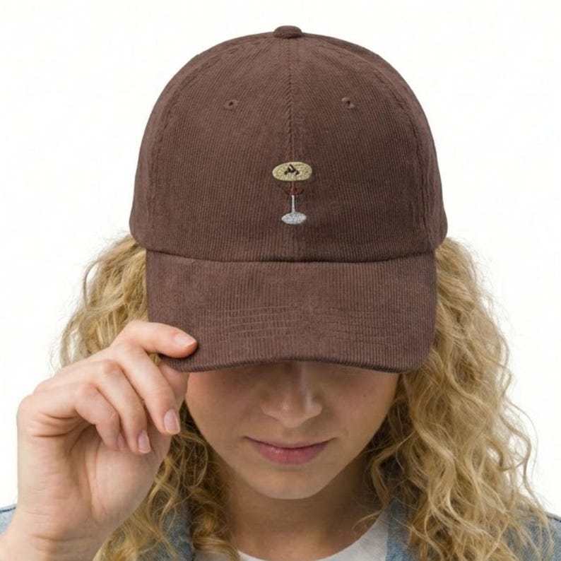 May include: A brown corduroy baseball cap with a small embroidered design of a cocktail glass. The cap has a curved brim and a button on top. The cap is being held by a person.