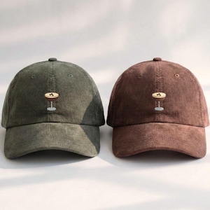 May include: Two corduroy baseball caps, one olive green and one brown. Each cap features an embroidered cocktail glass design. The caps have a curved brim and a button on top, suitable for casual wear.