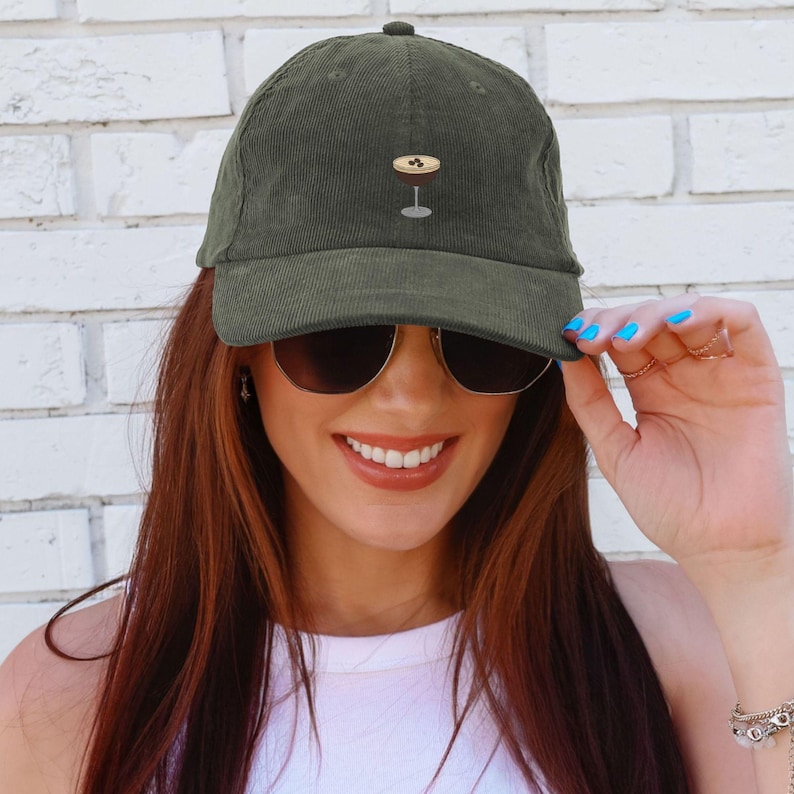 May include: Olive green corduroy baseball cap with a small embroidered cocktail glass design. The cap has a curved brim and a classic six-panel construction. The design is a simple, elegant accessory.
