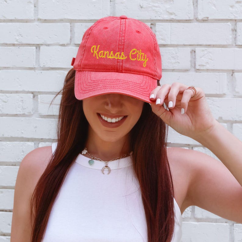 May include: A red baseball cap with the embroidered text "Kansas City" in yellow. The cap is worn by a person. The background is a white brick wall. The person is wearing a white tank top and a necklace.