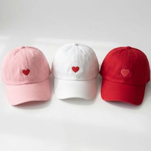 May include: Three baseball caps in pink, white, and red. Each cap has a curved peak and a small, embroidered red heart on the front. The caps are made of a soft fabric, suitable for casual wear.