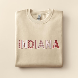 May include: A folded beige sweatshirt featuring the word "INDIANA" in a patchwork design. The letters are red and white, with patterns like stripes, polka dots, and plaid. The sweatshirt has a crew neck and a tag.