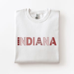 May include: A folded white sweatshirt with the word "INDIANA" across the front. The letters are in a red color with various patterns, including plaid, polka dots, and stripes. The sweatshirt is made of a soft material.