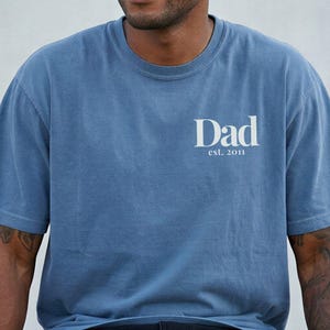 May include: A blue jean-colored t-shirt with the word "Dad" and the year "est. 2011" printed in white. The shirt is short-sleeved and has a crew neck. A black baseball cap is worn.