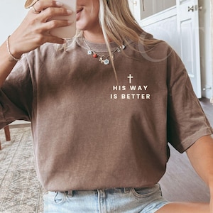May include: Espresso-colored t-shirt with the text "HIS WAY IS BETTER" and a cross graphic. The shirt is a relaxed fit and is paired with denim shorts and white sneakers. A straw hat is also visible.