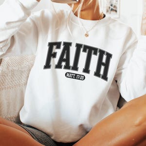 Puede incluir: Sudadera blanca con la palabra "FAITH" en letras negras, con "MATT. 17:20" debajo. La sudadera es de manga larga y tiene cuello redondo. La persona sostiene una taza negra.