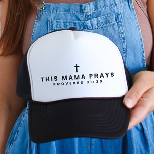 May include: A black and white trucker hat with the text "THIS MAMA PRAYS PROVERBS 31:30" and a cross symbol. The hat has a white front panel and a black brim and mesh back. The hat is held by a person wearing denim overalls.