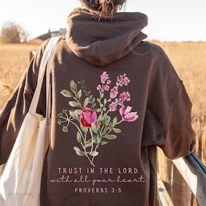 May include: Dark chocolate-colored hoodie with a floral design on the back, featuring pink and red flowers with green leaves. The text "TRUST IN THE LORD with all your heart PROVERBS 3:5" is printed below the flowers. A white tote bag is visible.