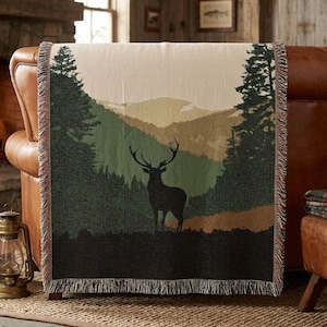 Op de afbeelding: Een geweven plaid met het donkere silhouet van een hert dat in een open plek in het bos staat. Het ontwerp bevat hoge dennenbomen aan de zijkanten, gelaagde bergketens in tinten groen en beige, en een gefranjerde rand aan de boven- en onderkant.