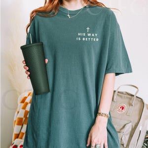 May include: Blue spruce-colored t-shirt with the text "HIS WAY IS BETTER" and a cross graphic. The shirt is a relaxed fit. A dark green textured tumbler is held in the left hand.