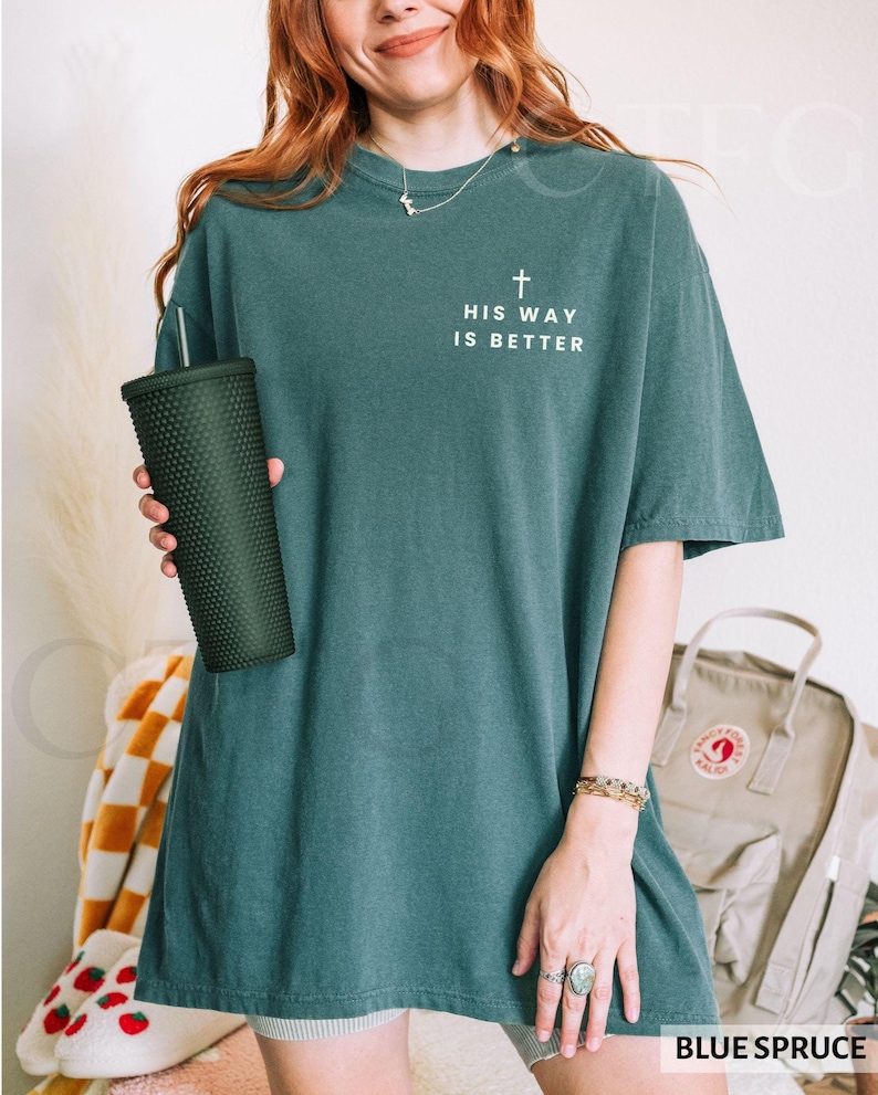 May include: Blue spruce-colored t-shirt with the text "HIS WAY IS BETTER" and a cross graphic. The shirt is a relaxed fit. A dark green textured tumbler is held in the left hand.