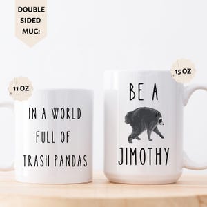 May include: Two white ceramic mugs with black text and a raccoon illustration. The left mug reads IN A WORLD FULL OF TRASH PANDAS. The right mug features a raccoon graphic above the text BE A JIMOTHY. Labels indicate 11 OZ and 15 OZ sizes.