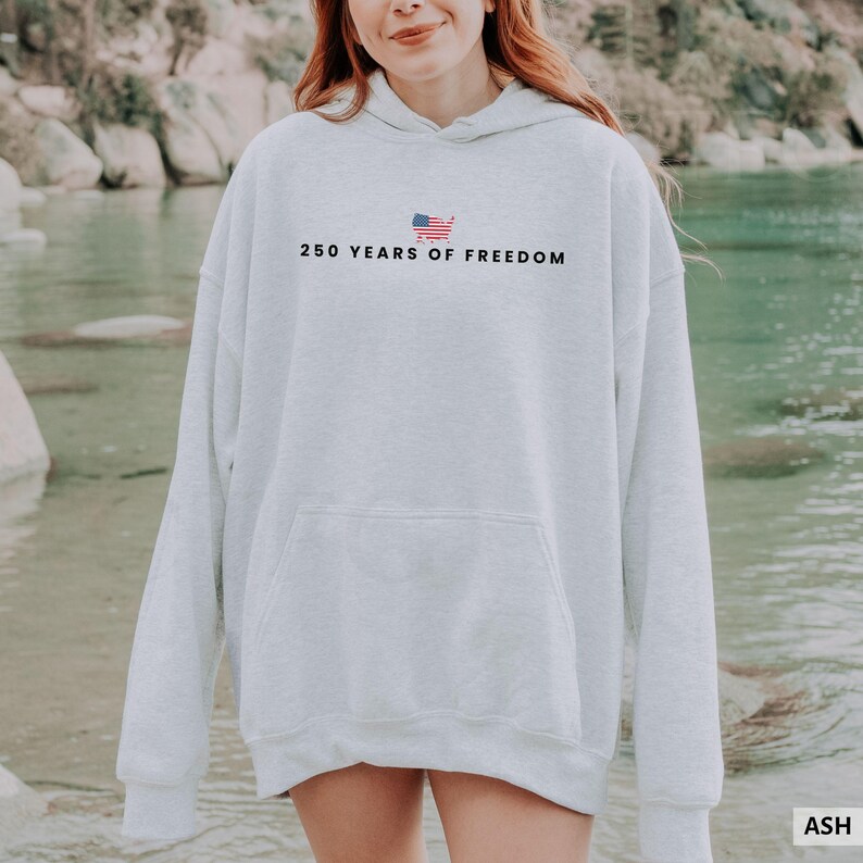 Puede incluir: Una sudadera con capucha gris claro con un bolsillo delantero y una capucha. El texto "250 YEARS OF FREEDOM" est&aacute; impreso en la parte delantera, encima de un peque&ntilde;o gr&aacute;fico de la bandera estadounidense. La sudadera est&aacute; hecha de un material suave.