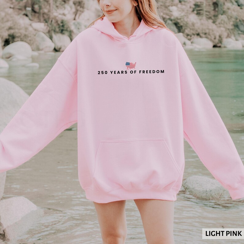 Puede incluir: Sudadera con capucha rosa claro con bolsillo delantero y capucha. El texto "250 YEARS OF FREEDOM" est&aacute; impreso en la parte delantera, junto con un peque&ntilde;o gr&aacute;fico de la bandera estadounidense. La sudadera es de color s&oacute;lido y est&aacute; hecha de un material suave.