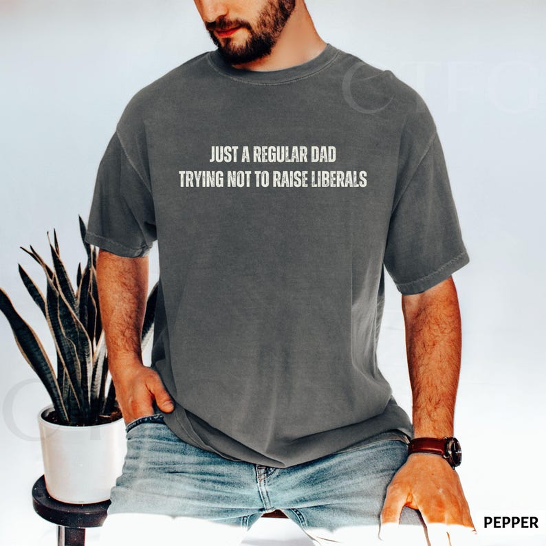 May include: A gray t-shirt with the text "JUST A REGULAR DAD TRYING NOT TO RAISE LIBERALS" in white. The shirt is worn by a person in blue jeans. The word "PEPPER" is in the bottom right corner.