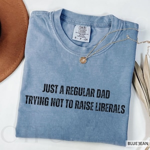 May include: A blue jean Comfort Colors t-shirt with the text "JUST A REGULAR DAD TRYING NOT TO RAISE LIBERALS" in black. The shirt has a crew neck and short sleeves. A gold necklace with a pendant is draped across the shirt.