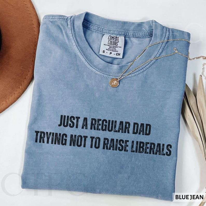 May include: A blue jean Comfort Colors t-shirt with the text "JUST A REGULAR DAD TRYING NOT TO RAISE LIBERALS" in black. The shirt has a crew neck and short sleeves. A gold necklace with a pendant is draped across the shirt.