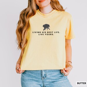 May include: A pale yellow short-sleeved t-shirt featuring a small graphic of a walking raccoon centered on the chest. Below the raccoon, the text reads in black capital letters: LIVING HIS BEST LIFE. LIVE YOURS. The shirt has a crew neckline.