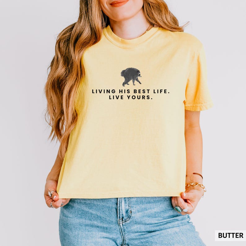 May include: A pale yellow short-sleeved t-shirt featuring a small graphic of a walking raccoon centered on the chest. Below the raccoon, the text reads in black capital letters: LIVING HIS BEST LIFE. LIVE YOURS. The shirt has a crew neckline.