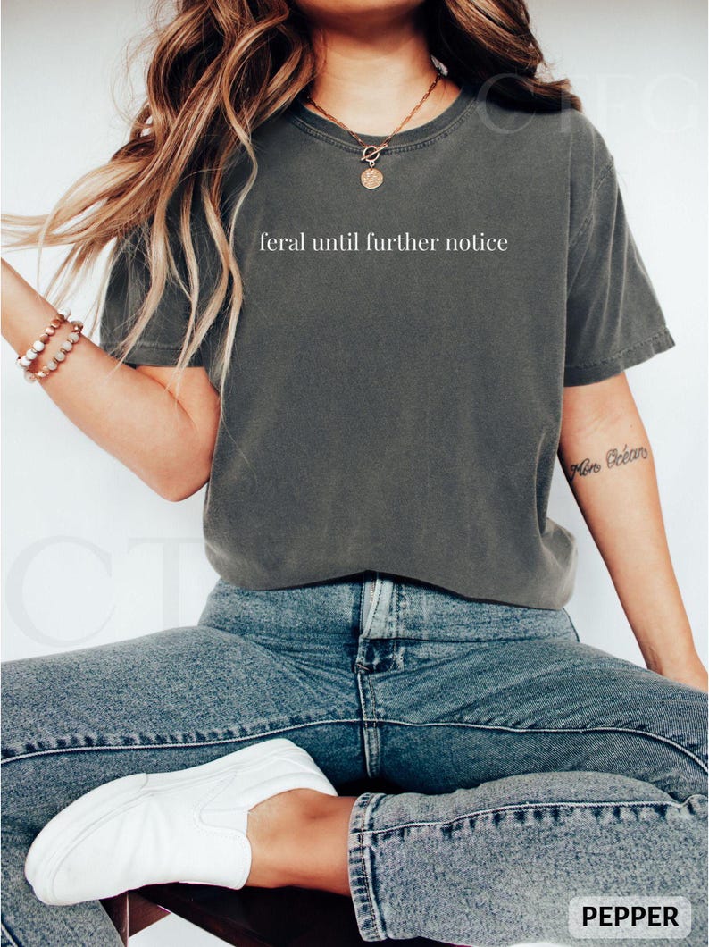 May include: A dark gray t-shirt with the text "feral until further notice" in white. The shirt is paired with blue jeans and white slip-on shoes. The word "PEPPER" is at the bottom right corner.