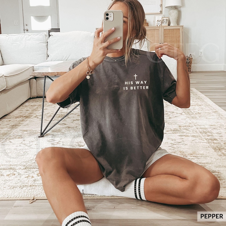 May include: Dark gray t-shirt with the text "HIS WAY IS BETTER" and a cross graphic. The shirt is a relaxed fit and has short sleeves. The model is wearing white shorts and black and white striped socks.