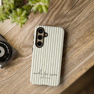 May include: A white and green striped phone case with the text "made for more" and "Ephesians 2:10". The phone case is on a wooden surface with a camera and greenery in the background.