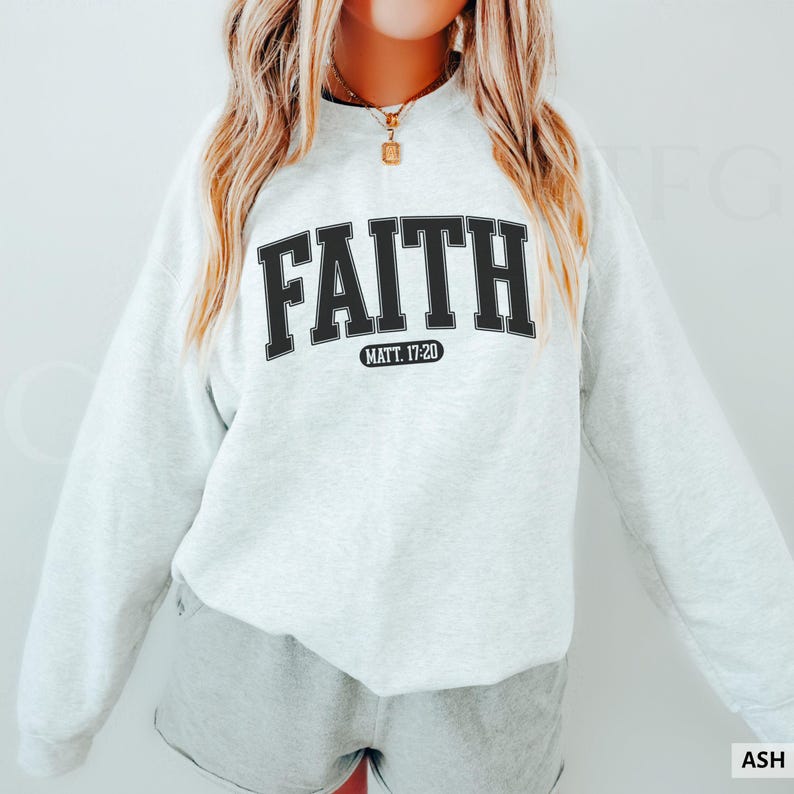 Puede incluir: Sudadera gris claro con la palabra "FAITH" en letras negras y gruesas en el pecho. Debajo, el texto "MATT. 17:20". La sudadera es de estilo casual.