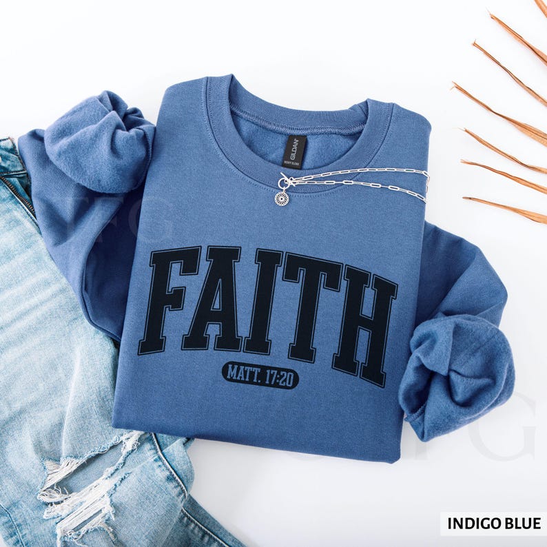 Puede incluir: Sudadera azul &iacute;ndigo con la palabra "FAITH" en letras negras. Debajo de la palabra "FAITH" est&aacute; el texto "MATT. 17:20". Un collar plateado con un colgante est&aacute; sobre la sudadera. La sudadera est&aacute; doblada sobre unos vaqueros azules.