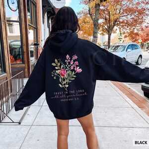 May include: Black hoodie with a floral design on the back, featuring pink and red flowers with green leaves. The text "TRUST IN THE LORD with all your heart PROVERBS 3:5" is printed below the flowers. The hoodie is a casual style.