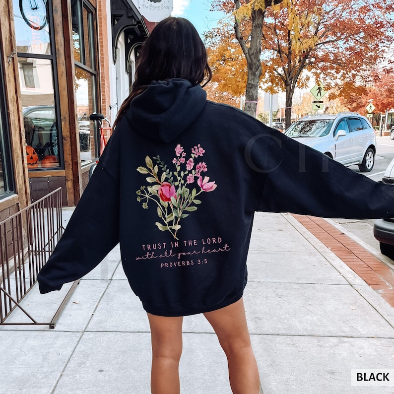 May include: Black hoodie with a floral design on the back, featuring pink and red flowers with green leaves. The text "TRUST IN THE LORD with all your heart PROVERBS 3:5" is printed below the flowers. The hoodie is a casual style.
