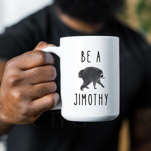 May include: A white ceramic mug featuring a black and white illustration of a raccoon in the center. Above the raccoon, the text reads BE A, and below the raccoon, the text reads JIMOTHY. The mug has a smooth finish and a curved handle on the side.