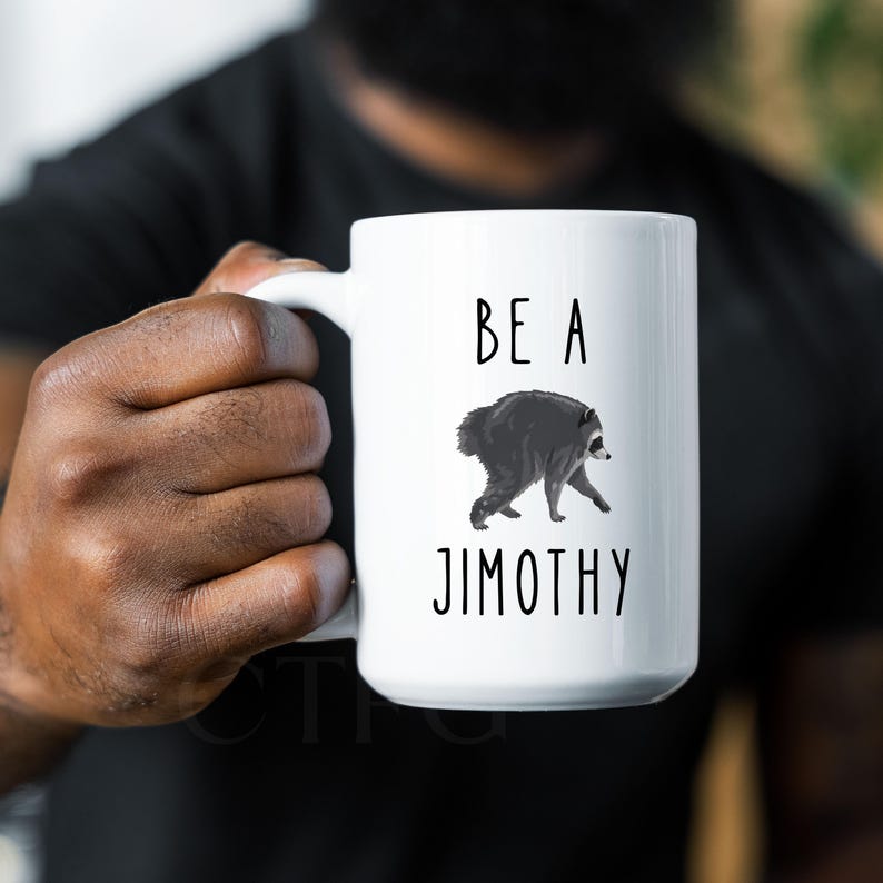 May include: A white ceramic mug featuring a black and white illustration of a raccoon in the center. Above the raccoon, the text reads BE A, and below the raccoon, the text reads JIMOTHY. The mug has a smooth finish and a curved handle on the side.