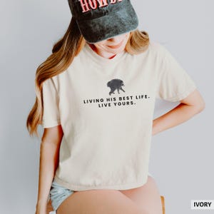 May include: An ivory t-shirt featuring a small graphic of a raccoon and the black text "LIVING HIS BEST LIFE. LIVE YOURS." The shirt has short sleeves and a crew neckline. A dark grey baseball cap with red lettering is worn above the shirt.